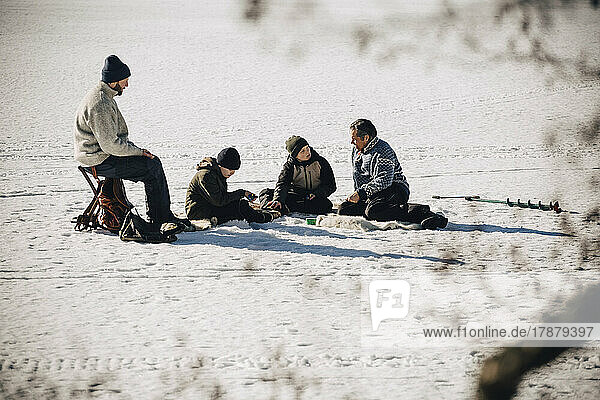 Mature men with sons sitting on snow while ice fishing during sunny day