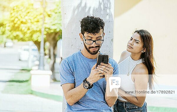 Suspicious girl spying on her boyfriend phone. Jealous girl spying on her boyfriend's cell phone in the park  Distrustful woman spying on her boyfriend's cell phone outdoors