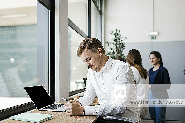 Lächelnder Geschäftsmann  der im Büro am Laptop sitzt und sein Mobiltelefon benutzt