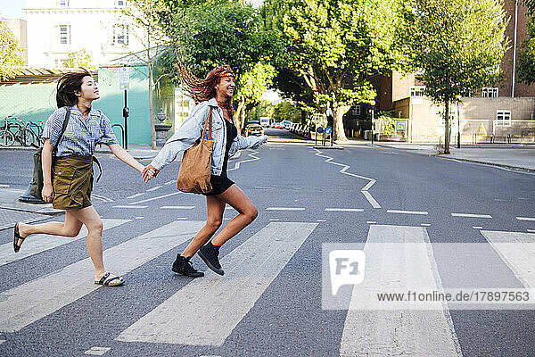 Cheerful friends holding hands and running on road in city