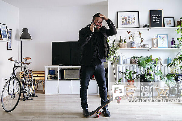Smiling businessman skateboard talking on smart phone in living room at home