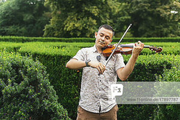 Young man with eyes closed playing violin by plants at park