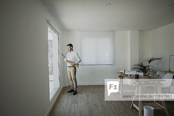 Architect holding rolled up blueprint using smart phone standing by window in office