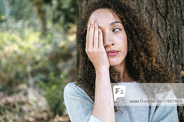 Young woman covering face with hand leaning on tree trunk at park