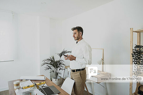 Thoughtful architect holding smart phone in office