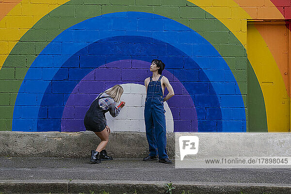 Young man wearing overalls standing by friend writing on rainbow mural