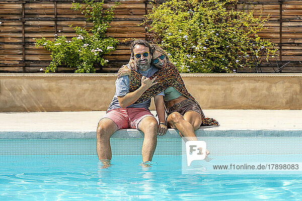 Happy mature woman hugging man sitting by pool on sunny day