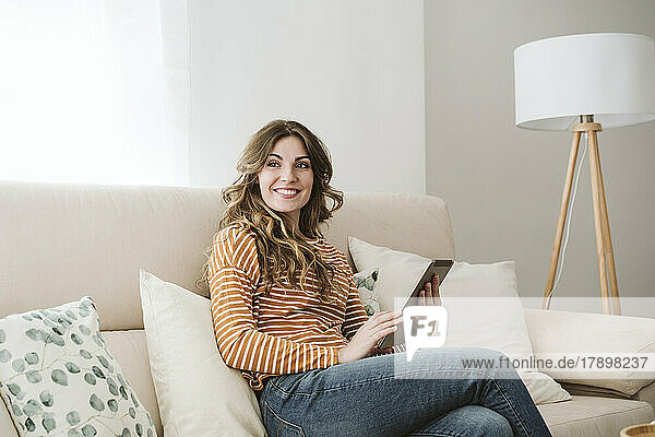 Lächelnde junge Frau hält digitales Tablet auf dem heimischen Sofa