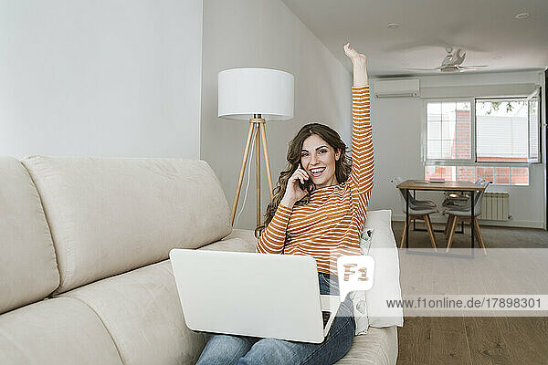 Happy young woman with laptop on sofa talking on the phone