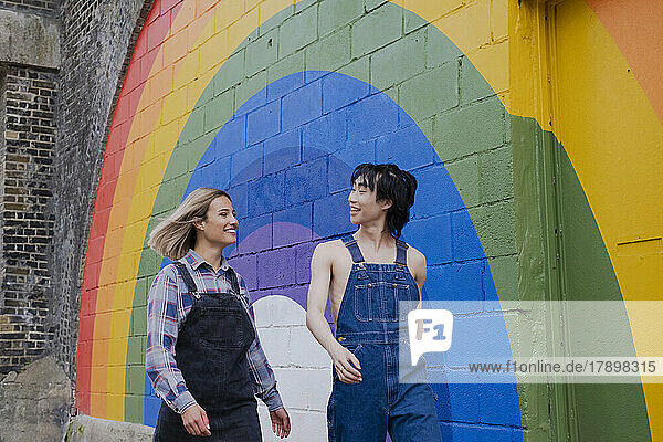 Fröhliche junge Frau und Mann gehen gemeinsam vor einem Regenbogenwandbild an der Wand spazieren