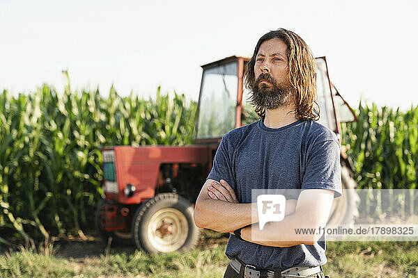 Contemplative bearded farmer with arms crossed standing in front of tractor