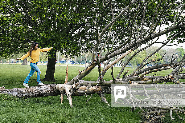 Junge Frau mit ausgestreckten Armen läuft auf umgestürztem Baum im Park