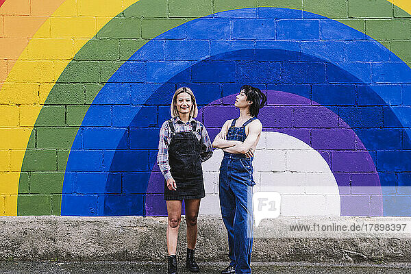 Happy young woman standing with friend in front of rainbow painted on wall