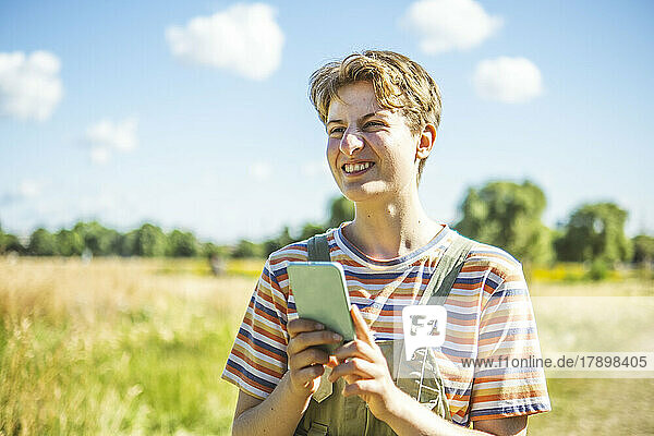 Smiling non-binary person with smart phone standing at park on sunny day