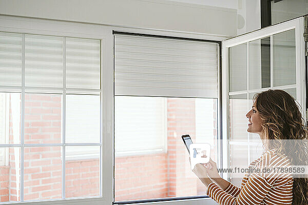 Young woman at home using mobile phone app to control blinds