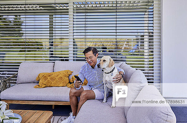 Smiling man using smart phone sitting with dog on sofa