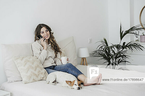 Young woman with coffee mug talking on phone sitting by dog in bedroom