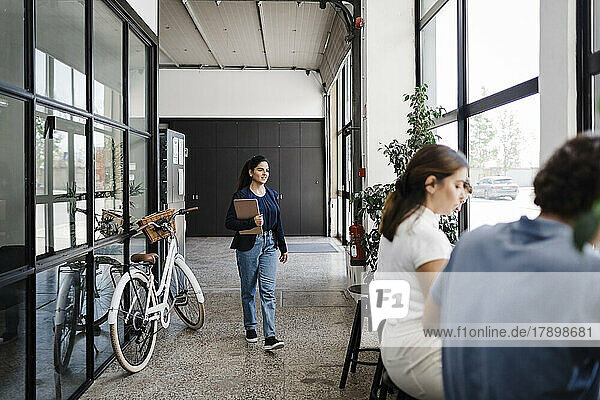 Young businesswoman with laptop walking by bicycle in corridor at office
