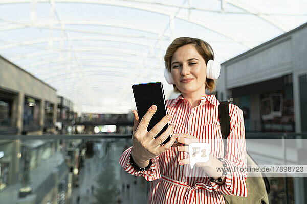 Smiling woman listening music through wireless headphones looking at mobile phone in shopping mall