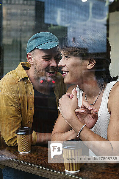 Smiling gay couple with coffee cups seen through window of cafe