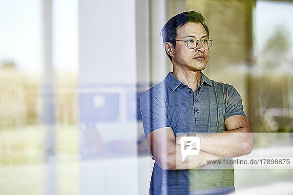Mature man standing with arms crossed seen through glass