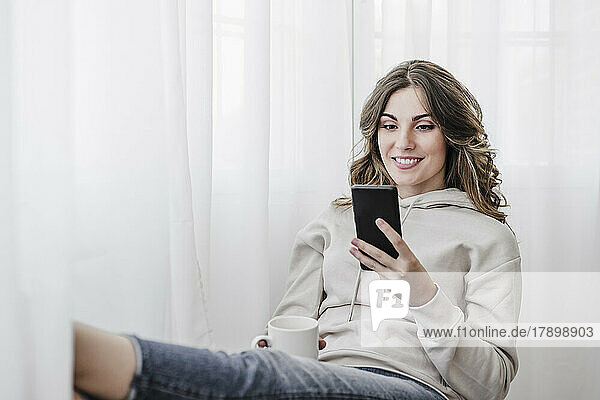 Smiling woman with cup using mobile phone at home