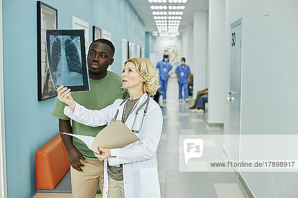 Mature doctor examining X-ray with patient standing in hospital corridor