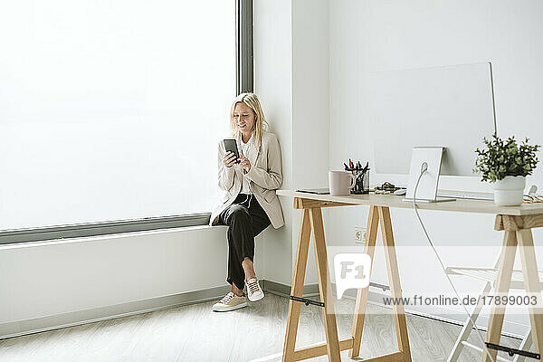 Businesswoman sitting on window sill in office holding mobile phone