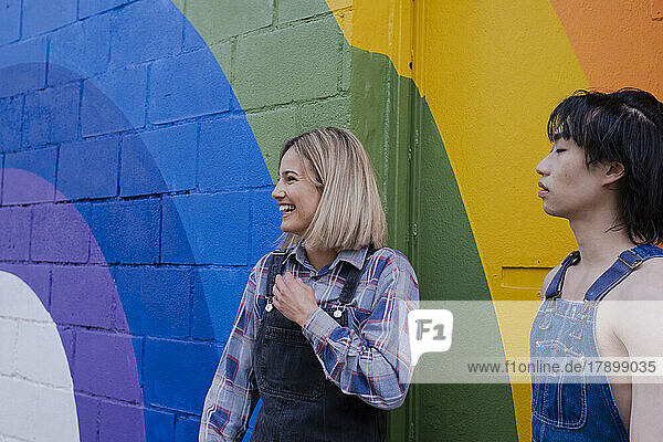 Happy young woman standing by friend in front of rainbow mural
