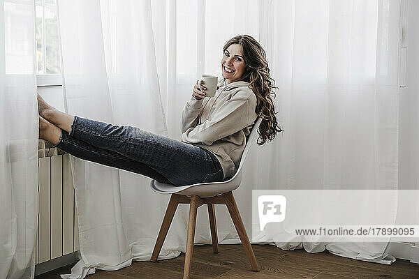 Happy woman with coffee mug sitting on chair at home