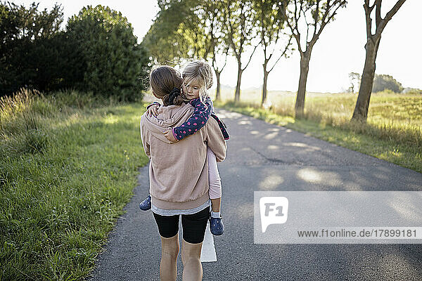 Mother carrying daughter walking on road by trees