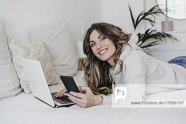 Happy woman with laptop and phone lying on bed at home