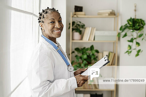Lächelnde Ärztin mit Klemmbrett am Fenster im Heimbüro