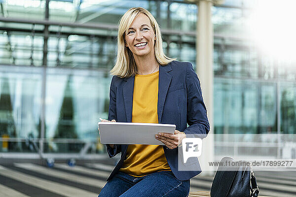 Smiling businesswoman with tablet PC sitting in front of building