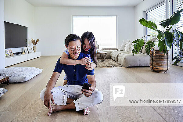 Happy woman with arm around man using smart phone on floor at home