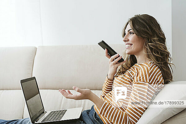 Young woman with laptop using mobile phone for a voicemail on sofa at home