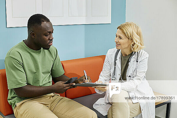 Patient unterschreibt Vertragspapier und sitzt mit Arzt auf der Couch in der Klinik