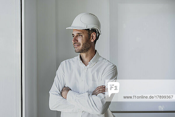 Smiling engineer with arms crossed looking through window in front of white wall