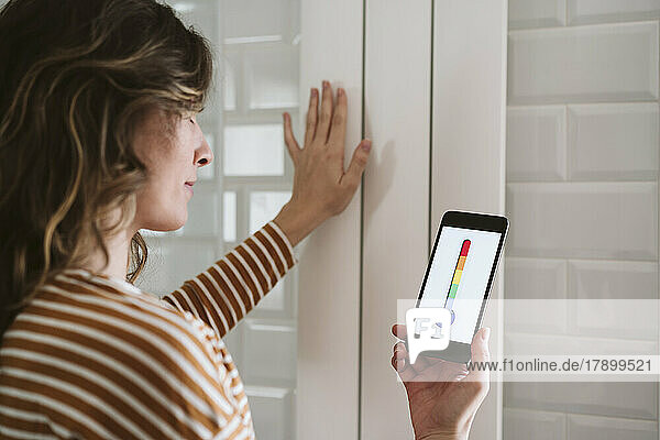 Young woman using smart phone app and touching radiator at home