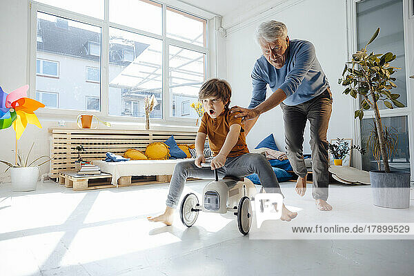 Cheerful grandfather pushing grandson sitting on toy car in living room