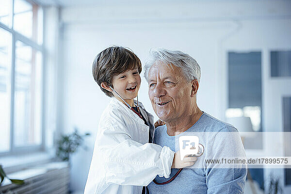 Fröhlicher Junge  der es als Arzt nachahmt und seinen Großvater zu Hause mit einem Stethoskop überprüft