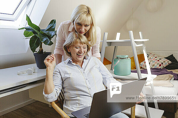 Blond woman with mother using laptop sitting on chair in bedroom