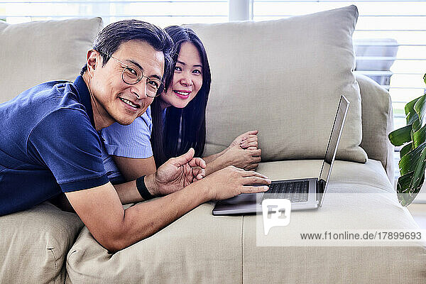 Happy mature couple with laptop lying on sofa at home