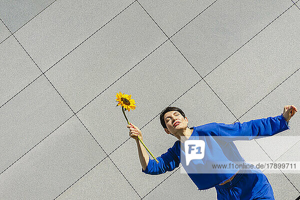 Woman with eyes closed holding sunflower in front of wall on sunny day