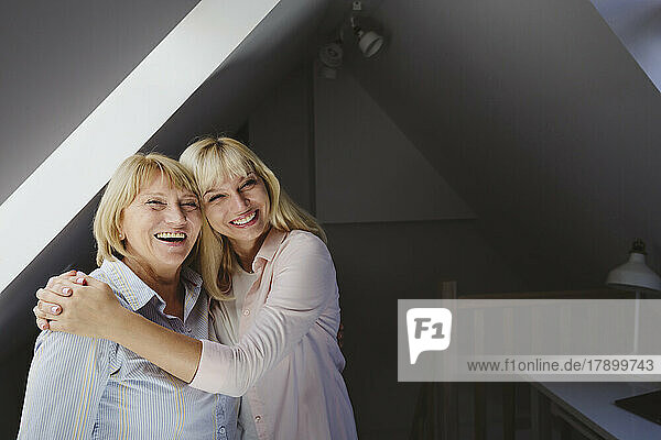 Cheerful blond woman hugging mother standing at home