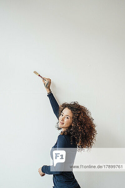 Lächelnde junge Frau mit erhobener Hand und Pinsel an weißer Wand