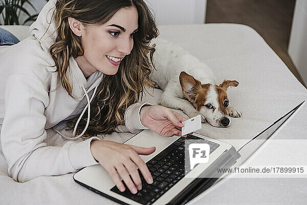 Young woman with credit card doing online shopping through laptop lying by dog at home