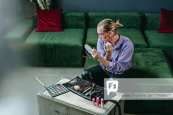 Man applying make-up looking in hand mirror sitting on sofa