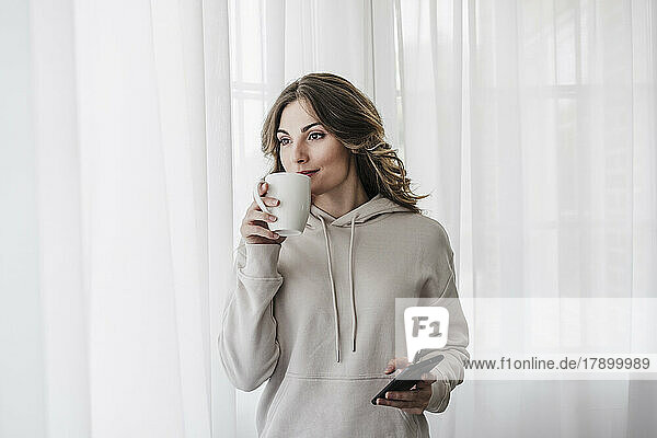 Frau mit Smartphone trinkt Kaffee vor weißem Vorhang zu Hause