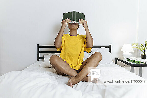 Woman covering her face with a book sitting cross-legged on bed